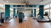 This single-floor fitness center is designed to support a comprehensive workout experience, combining thoughtful aesthetics with a full range of exercise equipment. The space is defined by striking teal accent walls paired with crisp white trim, creating a bold and energizing atmosphere. Warm hardwood-style flooring runs throughout, providing both visual warmth and a durable surface suited for a variety of training activities.

Large, multi-pane windows line the walls, flooding the interior with abundant natural light and offering views of the surrounding outdoor environment. The bright, open feel is further enhanced by recessed ceiling lighting and ceiling fans that maintain comfortable airflow during workouts.

The cardio section is well-appointed with multiple treadmills and elliptical machines, each equipped with individual display screens. The strength training area features a selection of selectorized weight machines, cable systems, and benches, providing options for both upper and lower body conditioning. A large wall-mounted flat-screen television is positioned for easy viewing from the strength training zone.

Full-length mirrors are strategically placed to visually expand the space and assist users in monitoring their form. The layout is open and uncluttered, allowing for ease of movement between equipment stations. The overall design balances functionality with a clean, modern aesthetic, making this fitness center an inviting and practical space for daily exercise.