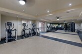 This fitness center is a comprehensively designed facility organized across a single level, offering distinct zones tailored to various workout modalities. The main cardio area is generously proportioned, housing a row of elliptical trainers and multiple treadmills arranged along the length of the room. The flooring throughout this zone transitions between rubberized dark-toned sections and lighter neutral surfaces, providing practical, durable support underfoot. Ceiling fans and recessed lighting maintain a bright, comfortable environment, while glass double doors along one wall admit natural light and offer views of an outdoor brick-paved courtyard.

The strength training area is positioned deeper within the facility and features a full rack of dumbbells, a cable machine, and additional cardio equipment, all reflected in large floor-to-ceiling mirrors that visually expand the space.

A dedicated studio room adjoins the main floor, finished with warm hardwood flooring, mirrored walls, and a coffered ceiling with skylights. This room is equipped with stability balls and open floor space suitable for yoga, stretching, or group classes. Sage green built-in cabinetry lines one wall, adding a refined, organized aesthetic.

Near the main entrance, a water fountain station, hand sanitizer dispenser, and a rack of kettlebells and medicine balls serve as practical amenities for facility users.
