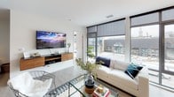 This single-floor residence showcases a contemporary design defined by clean lines, a neutral palette, and an abundance of natural light. The open-concept living area is anchored by expansive floor-to-ceiling windows with sleek dark frames, flooding the space with daylight and offering direct views of the surrounding urban streetscape. The living room comfortably accommodates a generous seating arrangement alongside a glass coffee table and hardwood-style flooring, creating a polished yet inviting atmosphere.

The kitchen features flat-front cabinetry in a rich dark wood finish, contrasted by a white subway tile backsplash and light quartz countertops. Stainless steel appliances, including a range, microwave, dishwasher, and refrigerator, provide a fully equipped cooking space. A peninsula with seating connects the kitchen to a dining area furnished with a warm wood table, ideal for casual meals.

The primary bedroom benefits from large windows offering urban views, soft carpeted flooring, and access to a walk-in closet with built-in shelving. A second bedroom features a distinctive circular window and its own walk-in closet. The primary bathroom includes a floating double vanity with an illuminated mirror and a soaking tub. An additional half bath completes the layout. Throughout, the design maintains a cohesive, modern sensibility.