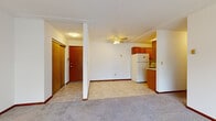 This single-floor unit is laid out to maximize functionality within a compact footprint. The main living area features neutral carpet flooring and white walls, creating a clean, versatile backdrop. A ceiling fan is centrally positioned overhead, and a structural column near the entryway adds architectural definition while subtly separating the entry from the living space. A closet with warm wood-toned sliding doors provides convenient storage near the entry.

The galley-style kitchen is efficiently arranged, featuring warm oak cabinetry on both walls, light tile flooring, a stainless sink with countertop workspace, and white appliances including a full-size refrigerator and dishwasher. Overhead cabinetry extends to the ceiling, offering substantial storage capacity.

The bedrooms are carpeted and feature white walls trimmed with rich wood-toned baseboards and window frames. Each room is served by baseboard heating, ensuring consistent warmth throughout. Large picture windows with bold wood frames bring in natural light and offer views of mature trees and the surrounding landscape. A sliding-door closet provides dedicated storage in one of the bedrooms. The 3D floor plan confirms a practical, well-organized arrangement of living, sleeping, kitchen, and bathroom spaces throughout the unit.