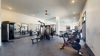 Located at 1875 Maple Dr in Kennesaw, GA, this well-appointed fitness center offers a generously sized, single-floor layout designed to accommodate a wide range of workout needs. The open floor plan is covered in durable rubber flooring throughout, providing a safe and comfortable surface for all types of exercise. The space is filled with an extensive array of equipment, including multi-station cable machines, power racks, weight benches, and a dedicated free weights area. A row of treadmills lines the perimeter wall, positioned in front of large floor-to-ceiling windows and glass doors that flood the space with natural light and offer views of the outdoor surroundings. Spinning bikes with interactive displays occupy a dedicated zone near one of the glass-door entrances, while a functional training area features a wall-mounted rack stocked with kettlebells, medicine balls, and a punching bag. Full-length mirrors along the walls enhance the sense of openness and help users monitor their form. Recessed lighting and modern pendant ceiling fans provide even illumination and air circulation throughout. Structural columns are neatly integrated into the layout, and clean white walls contribute to the bright, contemporary aesthetic of this well-equipped community fitness facility.