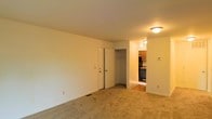 This single-floor space presents a straightforward and functional layout. The main living area is finished with wall-to-wall beige carpeting, which lends a warm, neutral tone throughout. Walls are painted in a clean off-white, keeping the space feeling open and easy to personalize. Overhead lighting is provided by a flush-mount ceiling fixture, offering practical illumination.

A closet is integrated directly into the living area, featuring a standard bi-fold door and a visible hanging rod, providing convenient built-in storage. Adjacent to the closet, an opening leads directly into the kitchen, which is partially visible from the main room. The kitchen features overhead wood-toned cabinetry, dark appliances including what appears to be an oven range, and a hard-surface floor that contrasts with the carpeted living space. A small countertop work area is also visible.

The overall layout connects the living and kitchen areas in a compact, efficient manner. Neutral finishes throughout make the space adaptable, while the inclusion of built-in storage and a functional kitchen provide essential everyday conveniences. The space is well-suited for straightforward, comfortable living.