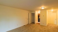 This single-floor space presents a straightforward and functional layout. The main living area is finished with wall-to-wall beige carpeting, which lends a warm, neutral tone throughout. Walls are painted in a clean off-white, keeping the space feeling open and easy to personalize. Overhead lighting is provided by a flush-mount ceiling fixture, offering practical illumination.

A closet is integrated directly into the living area, featuring a standard bi-fold door and a visible hanging rod, providing convenient built-in storage. Adjacent to the closet, an opening leads directly into the kitchen, which is partially visible from the main room. The kitchen features overhead wood-toned cabinetry, dark appliances including what appears to be an oven range, and a hard-surface floor that contrasts with the carpeted living space. A small countertop work area is also visible.

The overall layout connects the living and kitchen areas in a compact, efficient manner. Neutral finishes throughout make the space adaptable, while the inclusion of built-in storage and a functional kitchen provide essential everyday conveniences. The space is well-suited for straightforward, comfortable living.