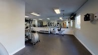 Located at 6300 Pewter Ave in North Chesterfield, VA, this single-floor fitness center is thoughtfully designed to support a full range of workout routines. The space is organized into distinct functional zones, beginning with a welcoming entry area that features durable rubber flooring, wall-mounted sanitation dispensers, a wooden bench for changing, and direct access to a clean restroom equipped with a vanity, mirror, and tiled flooring.

The main exercise floor is well-lit by recessed flush-mount ceiling fixtures that illuminate the entire workout area evenly. Full-length mirrors line the walls, visually expanding the space and allowing users to monitor their form during exercise. The cardio zone is equipped with multiple treadmills, elliptical trainers, and a recumbent bike, arranged to maximize open floor space. A wall-mounted television provides entertainment during workouts.

The strength training area features a multi-function cable machine, a weight bench, and a organized dumbbell rack stocked with a range of free weights. Decorative wall art adds a polished, modern aesthetic to the neutral gray-toned walls. The rubber-tiled flooring throughout provides both safety and durability. Natural light filters in through windows, complementing the overhead lighting to create a bright and motivating environment suitable for daily fitness use.