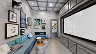 This dedicated theater space is thoughtfully designed to deliver an immersive entertainment experience without sacrificing style. The room is anchored by a large projection screen mounted within a sleek, built-in gray cabinetry unit that spans the far wall, providing both a polished focal point and integrated media storage. Overhead, a striking coffered ceiling with recessed lighting adds architectural depth and a refined aesthetic while contributing to the room's acoustics. The seating arrangement features a spacious teal sectional sofa that lines two walls, offering generous, comfortable viewing capacity. A set of industrial-style drum ottomans with metallic tops serve as flexible accent pieces and additional seating in the center of the room. The walls are adorned with a curated gallery of framed artwork, lending the space a creative and personalized atmosphere. Neutral gray tones throughout — from the walls and cabinetry to the polished concrete-style flooring — create a cohesive, contemporary palette. A glass-paneled interior door adds a sophisticated touch while maintaining visual openness. In-ceiling speakers are also visible, pointing to a fully integrated audio-visual setup built for a true cinematic experience.