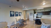This single-story space showcases an open-concept layout that seamlessly connects the dining area and kitchen, creating a cohesive and functional living environment. The dining area features hardwood-style flooring and is furnished with a round wooden table surrounded by mixed-style chairs, adding an eclectic yet coordinated aesthetic. A series of three botanical wall art pieces in navy blue tones anchor the dining space, providing a striking focal point against the neutral, warm-toned walls.

Overhead, a combination of track lighting and a ceiling fan ensures both practical illumination and comfort throughout the space. The kitchen, visible from the dining area, is equipped with dark cabinetry, a stainless steel refrigerator, and a light-colored countertop island with bar-height seating. A pendant light hangs above the kitchen area, adding a decorative touch. A topiary plant on the counter introduces a natural element to the kitchen's design.

The open sightlines between the two areas enhance the sense of spaciousness, while neutral wall tones and consistent flooring unify the overall aesthetic. A hallway with a white paneled door leads toward additional areas of the space, suggesting further rooms beyond what is visible.