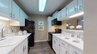 This single-story residence offers a well-organized layout that balances functionality and comfort throughout its connected living spaces. The galley-style kitchen is a standout feature, showcasing crisp white cabinetry with brushed silver hardware extending from floor to ceiling, providing generous storage. Light-toned countertops contrast neatly with black appliances, including an electric range, over-the-range microwave, and refrigerator. Wood-look flooring runs throughout the kitchen and continues into the adjacent dining area, which is illuminated by a pendant light and features a pass-through opening connecting it to the kitchen. The living room benefits from natural light streaming through a set of French doors that open to an outdoor space, creating a bright and welcoming atmosphere. Two bedrooms are finished with plush carpeting and ceiling fans; each room offers closet storage and windows with outdoor views. The full bathroom includes a tub-shower combination, a white vanity with ample counter space, and wood-look flooring that ties cohesively to the rest of the unit. Neutral warm-beige wall tones throughout the residence create a calm, cohesive ambiance.