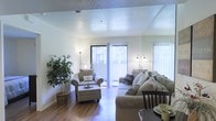 This single-floor space offers a welcoming and well-lit interior designed for comfortable everyday living. The main living area is anchored by warm hardwood flooring that extends throughout, lending a sense of warmth and continuity to the space. Large sliding glass doors allow abundant natural light to pour in, complemented by vertical blinds that offer flexible light control and privacy. The layout feels open and airy, with room for both a generously sized sectional sofa and a cushioned armchair with an ottoman, creating a relaxed and functional seating arrangement. A decorative indoor plant adds a natural, organic touch to the corner of the room, enhancing the overall ambiance. Soft, neutral wall tones keep the space feeling clean and calm, while floor lamps provide warm supplemental lighting for the evenings. A dining area is partially visible, suggesting a natural flow between living and dining spaces. The textured ceiling adds a subtle architectural detail overhead. Overall, the space presents a bright, tidy, and inviting atmosphere well-suited for both relaxation and everyday entertaining.