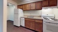 This single-floor, 2-bedroom, 1-bathroom unit offers a practical and straightforward layout designed for comfortable everyday living. The living area features a combination of plush carpeting and hardwood-style flooring, with a ceiling fan providing overhead airflow and lighting. A half-wall divides this space from the adjacent kitchen, creating an open yet defined flow between the two areas. Natural light enters through a large window, and a wall-mounted air conditioning unit provides supplemental cooling alongside baseboard heating.

The kitchen is equipped with warm-toned wood cabinetry extending to the upper and lower sections of the walls, offering generous storage space. A stainless steel sink sits beneath the cabinetry, complemented by laminate countertops. White appliances including a refrigerator, gas range, and dishwasher are included. The kitchen features durable dark hardwood-style flooring.

Both bedrooms are carpeted and neutrally painted, with windows offering views of outdoor greenery and baseboard heating along the walls. The full bathroom includes a tiled tub-shower combination with chrome fixtures. Throughout the unit, neutral wall tones and clean finishes create a simple, move-in-ready environment.