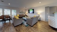 This single-floor communal space is thoughtfully designed to foster comfort and connection. The open floor plan flows seamlessly across rich hardwood-style flooring, creating a cohesive and inviting atmosphere throughout. The main lounge area is generously sized, accommodating multiple distinct seating arrangements furnished with plush gray sofas and armchairs accented by vibrant yellow throw pillows and layered area rugs. Wall-mounted flat-screen televisions are positioned for easy viewing from the various seating groupings, making the space well-suited for both relaxed gatherings and community programming. A cozy alcove hosts a pair of light blue swivel chairs alongside a side table and floor lamp, offering a quieter nook for conversation or reading. The kitchenette area is equipped with white shaker-style cabinetry, dark countertops, a coffee and beverage station, a compact refrigerator, and a sink with a gold-toned faucet. Large windows with white horizontal blinds line the kitchenette, flooding the space with natural light and offering pleasant outdoor views. Recessed lighting and carefully placed lamps throughout contribute to a warm, well-lit ambiance. The neutral gray walls and clean architectural lines give the space a modern yet approachable character, making it an ideal setting for community interaction and relaxation.