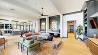 This thoughtfully designed clubhouse in Baton Rouge, Louisiana, offers a welcoming and versatile gathering space spread across a single floor. The open-concept layout connects multiple functional areas seamlessly, all unified by light hardwood flooring, crisp white walls, and bold dark-trimmed architectural details that lend a sophisticated, contemporary aesthetic.

The lounge area serves as the heart of the space, anchored by a large sectional sofa, plush green accent chairs, and a patterned area rug. Dark teal paneled alcoves with built-in desks provide practical workstations, while a wall-mounted TV flanked by dark built-in shelving creates a media and entertainment focal point. A ceiling fan overhead and an abundance of natural light pouring through oversized, dark-trimmed windows and French doors enhance the airy ambiance throughout.

The kitchen is a standout feature, boasting sage green cabinetry with brushed gold hardware, white quartz countertops, a herringbone tile backsplash, open floating shelves, and a large central island. Striking gold dome pendant lights add a warm, refined touch. A dining area adjacent to the kitchen accommodates a long communal table with vibrant blue chairs beneath matching gold pendants. The space also includes a game area with a foosball table, completing its appeal as a dynamic, multi-use community hub.