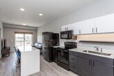 This single-floor unit showcases a contemporary design with a clean, neutral color palette and light wood-style flooring carried throughout. The open-concept kitchen is a standout feature, equipped with a full suite of black appliances — including a French door refrigerator, electric range, over-the-range microwave, and dishwasher. Two-tone cabinetry pairs white upper cabinets with dark lower cabinets, complemented by light countertops. A kitchen island with seating for multiple provides additional workspace and a casual dining option.

The living area is well-proportioned and bathed in natural light from sliding glass doors that open to a private balcony. The space offers a comfortable layout suitable for both relaxing and entertaining. The bedroom is thoughtfully sized, featuring a window with exterior views and room for a bed and dedicated study area.

Two bathrooms serve the unit, each consistently finished with dark vanity cabinetry, white countertops, framed mirrors, and a shower-tub combination. Recessed lighting throughout and a smart thermostat contribute to the unit's modern, functional character.