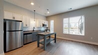 Located at 750 E Broad St in Columbus, Ohio, this single-floor unit offers a thoughtfully designed layout with modern finishes throughout. The kitchen is a standout feature, equipped with stainless steel appliances including a refrigerator, stove, microwave, and dishwasher. White shaker-style upper cabinets contrast with gray lower cabinets, complemented by a subway tile backsplash and dark countertops. Pendant lighting and a freestanding island add both functionality and style, while a large window frames views of the Columbus skyline, filling the space with natural light. Durable wood-style flooring runs throughout the unit, lending a warm, cohesive feel to each room. A well-proportioned hallway connects the living areas to additional rooms, with a neatly tucked in-unit stacked washer and dryer providing added convenience. The bedroom features soaring ceilings that enhance the sense of openness, with direct access to the bathroom. The bathroom is finished with deep teal accent walls, a glass-enclosed walk-in shower with black hardware, and light tile flooring, creating a bold yet clean aesthetic. The overall ambiance is contemporary and functional, ideal for urban living in the heart of Columbus.