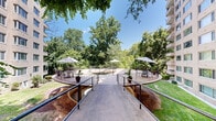 Located at 3640 16th St NW in Washington, DC, this two-level outdoor courtyard is a thoughtfully designed communal space set against the backdrop of a multi-story building with a classic light brick facade. The space is defined by its striking circular sunken area at the courtyard's center, enclosed by a curved metal railing and accessible via descending steps — a distinctive architectural focal point that draws the eye and anchors the overall layout.

Spanning two levels, the courtyard is connected by gently sloping ramps with metal handrails, ensuring accessibility throughout. The upper and lower terraces are finished with aggregate paving and are outfitted with metal bistro-style seating arrangements shaded by large patio umbrellas, creating inviting areas for outdoor gathering and relaxation.

Lush, mature trees surround the perimeter of the courtyard, providing ample shade and a sense of natural enclosure that softens the building's architectural presence. Potted tropical plants are strategically placed throughout, adding pops of greenery and enhancing the garden-like atmosphere.

The space benefits from abundant natural light, with open sightlines to the sky and the surrounding tree canopy. The overall ambiance is serene and well-maintained, offering a tranquil urban retreat within the heart of Washington, DC's 16th Street corridor.