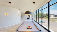 Located at 1500 SW Aileron Cir in Bentonville, Arkansas, this single-floor amenity space offers a thoughtfully designed environment centered around recreation and entertainment. The layout is divided into two distinct zones, each with its own character and purpose.

The game lounge is a bright, open area defined by soaring ceilings and an expansive wall of floor-to-ceiling windows that flood the space with natural light and provide sweeping outdoor views of the surrounding community. The patterned, textured carpeting in white and gold tones adds warmth underfoot, while bold dome pendant light fixtures anchor the room visually. Multiple billiard tables are positioned throughout the open layout, complemented by a stylish built-in cabinetry wall with a mounted television — ideal for social gatherings. Curved architectural walls lend a modern, fluid quality to the design.

In contrast, the private theater room delivers an immersive cinematic experience. The room is finished in deep, dark tones with a black ceiling and muted walls that eliminate distractions and enhance screen visibility. Rows of plush leather recliner seats are arranged on tiered platforms, ensuring clear sightlines to the large projection screen. Recessed ceiling lighting provides subtle ambiance without disrupting the viewing experience. Together, these two spaces form a compelling and versatile entertainment destination.