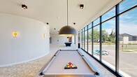 Located at 1500 SW Aileron Cir in Bentonville, Arkansas, this single-floor amenity space offers a thoughtfully designed environment centered around recreation and entertainment. The layout is divided into two distinct zones, each with its own character and purpose.

The game lounge is a bright, open area defined by soaring ceilings and an expansive wall of floor-to-ceiling windows that flood the space with natural light and provide sweeping outdoor views of the surrounding community. The patterned, textured carpeting in white and gold tones adds warmth underfoot, while bold dome pendant light fixtures anchor the room visually. Multiple billiard tables are positioned throughout the open layout, complemented by a stylish built-in cabinetry wall with a mounted television — ideal for social gatherings. Curved architectural walls lend a modern, fluid quality to the design.

In contrast, the private theater room delivers an immersive cinematic experience. The room is finished in deep, dark tones with a black ceiling and muted walls that eliminate distractions and enhance screen visibility. Rows of plush leather recliner seats are arranged on tiered platforms, ensuring clear sightlines to the large projection screen. Recessed ceiling lighting provides subtle ambiance without disrupting the viewing experience. Together, these two spaces form a compelling and versatile entertainment destination.