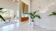 This elegant single-floor club lobby offers a generously proportioned, open-plan layout designed to impress and welcome. The space is anchored by dramatic soaring ceilings that amplify the sense of volume and airiness throughout. Polished terrazzo flooring flows seamlessly across the entire area, reflecting natural light that pours in through oversized floor-to-ceiling windows framing lush tropical views of palm trees and the surrounding streetscape.

At the entrance, a formal reception desk finished in warm wood tones stands against a striking travertine-clad feature wall, illuminated by a backlit branded sign that serves as the focal point of arrival. Tall tropical palms and bird-of-paradise plants in sleek white planters are positioned throughout, reinforcing a resort-inspired aesthetic.

The lounge area offers a refined seating arrangement with a mid-century modern sofa, accent chairs, and a sculptural wooden coffee table, all centered on a textured area rug. Pendant globe lights hang above, adding warmth and dimension to the bright interior.

A casual café-style refreshment station with a wood-paneled counter and high communal table provides a social gathering point. Adjacent to this, a dedicated meeting nook features a round tulip-style table, upholstered chairs, and teal accent walls with fluted wood paneling, creating an intimate yet design-forward workspace. Curated artwork adds personality throughout the space.
