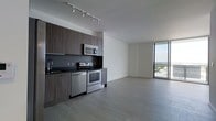 This contemporary single-floor unit showcases a clean, open-concept layout designed with modern living in mind. The kitchen is outfitted with dark wood cabinetry, dark countertops, and a contrasting backsplash that adds depth to the space. Stainless steel appliances — including a range, microwave, and dishwasher — complement the kitchen's polished aesthetic, while a track lighting system overhead ensures the workspace is well-lit. The kitchen flows seamlessly into a generous living area, creating an inviting and functional open-plan environment. Light-toned wood-style flooring runs throughout, lending warmth and visual continuity to the space. A standout feature is the floor-to-ceiling window wall at the far end of the living area, which floods the interior with abundant natural light and frames sweeping views of the surrounding cityscape and landscape beyond. The overall ambiance is sleek and minimalist, with clean lines, neutral tones, and an uncluttered design that highlights the architectural quality of the space. The combination of modern finishes, natural light, and panoramic views makes this a bright, sophisticated, and highly functional living environment.