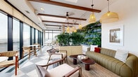Located at 519 S Washington Ave in Minneapolis, this thoughtfully designed conservatory lounge offers a refined and nature-inspired retreat. The single-floor space is defined by its striking floor-to-ceiling glass walls, which flood the interior with natural light and frame sweeping panoramic views of the Minneapolis skyline. A bold checkered stone tile floor anchors the layout, complementing the warm, organic aesthetic throughout.

A dramatic living green wall serves as a central design feature, layered with cascading foliage that brings a vibrant, botanical energy to the lounge. Lush freestanding planters and tropical plants are thoughtfully positioned throughout the space, reinforcing its conservatory identity. Exposed wood ceiling beams and woven rattan pendant lights add warmth and texture, while ceiling fans contribute to the relaxed, airy atmosphere.

The lounge accommodates multiple seating arrangements, including a striped olive-green sofa, neutral sectional seating with colorful accent pillows, and mid-century-style wooden armchairs. A sleek built-in beverage station with white subway tile backsplash and dark countertops provides a functional amenity. A standout feature is a white grand piano positioned beside the expansive windows, offering an elegant focal point with a city backdrop. Intimate bistro-style seating near the glass walls creates an ideal setting for taking in the urban views.