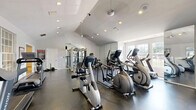 Located at Lakeshore Trail East in Indianapolis, Indiana, this fitness center spans a single floor and is divided into two distinct exercise rooms, each thoughtfully designed to support a full range of workout activities.

The first room is characterized by a warm, inviting atmosphere with a striking natural wood ceiling and bold blue accent walls. Large wall mirrors expand the sense of space, while a wall-mounted flat-screen TV adds entertainment value. This room is stocked with a comprehensive free weight rack, medicine balls, stability balls, a weight bench, and a stationary bike, making it ideal for strength and functional training. Durable flooring provides a stable surface throughout.

The second, larger exercise room features soaring vaulted ceilings with recessed lighting, a ceiling fan, and abundant natural light streaming through multiple windows with views to the outdoors. Floor-to-ceiling mirrors line one wall, amplifying the open, airy feel of the space. This room houses a full selection of cardio machines, including treadmills, ellipticals, a recumbent bike, and a multi-station cable machine, catering to a wide variety of fitness routines. The dark rubberized flooring contrasts cleanly with the bright white walls and ceiling.

Together, the two rooms create a comprehensive and well-appointed fitness facility suited for both cardio and strength training.