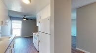 Located at 443 Gateway Dr in Pacifica, CA, this single-story unit offers a clean, functional layout with consistent wood-style flooring and neutral gray wall tones throughout. The galley-style kitchen is well-appointed with white cabinetry extending to the ceiling for ample storage, dark countertops lining both sides, a white electric range, refrigerator, and an under-cabinet sink. Recessed lighting illuminates the kitchen corridor, which connects conveniently to the main living areas. The living room is a generously sized, open space featuring a textured ceiling, a ceiling fan with light fixture, and a sliding glass door that opens to a private balcony with outdoor views, bringing in natural light. Adjacent rooms provide flexible living arrangements, with each space finished in the same cohesive flooring and neutral palette. A bedroom features mirrored sliding closet doors offering practical storage. The bathroom is straightforward and clean, equipped with a tub-shower combination, chrome fixtures, and a grab bar. Throughout the unit, white baseboards provide crisp definition, contributing to a tidy, move-in-ready ambiance suited to everyday living on the California coast.