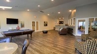 This single-floor communal space is thoughtfully designed to provide a welcoming and versatile environment for recreation and socializing. The open-concept layout is anchored by rich hardwood-style flooring that runs throughout, lending warmth and cohesion to the entire space. Vaulted ceilings rise overhead, enhancing the sense of openness and allowing recessed lighting to illuminate the room evenly.

The recreation area takes center stage with a classic pool table, offering an inviting focal point for entertainment. Adjacent to it, a comfortable lounge seating arrangement — featuring a plush sofa with accent pillows and a round ottoman on a area rug — creates an ideal spot for conversation and relaxation. Colorful wall art adds personality and visual interest throughout the space.

Toward the back, a stylish bar counter with pendant lighting and bar stool seating serves as a social hub, complemented by a pass-through window that connects to an adjacent service area. A set of French doors provides access to a separate room, adding functional versatility to the layout.

Overall, the space strikes a balance between casual comfort and modern design, making it well-suited for gatherings, leisure activities, and everyday community use.