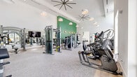 Located along the Tamiami Trail in Naples, Florida, this contemporary fitness center is designed to deliver a comprehensive workout experience in a bright, well-appointed setting. The space is characterized by soaring double-height ceilings that create an open, airy atmosphere, complemented by large floor-to-ceiling windows and clerestory glazing that flood the interior with abundant natural light and offer views of the surrounding outdoor amenities, including a pool area.

The main workout floor is finished with plush, light-toned carpet and features an extensive selection of Matrix-brand strength training machines, including cable systems, leg press, and multi-functional units. A dedicated free weights section with dumbbells and a heavy bag rack serves users seeking functional and combat training. Full-length mirrors expand the sense of space throughout the strength zone, which is accented by a bold green feature wall with decorative gold sconces.

The cardio zone is generously equipped with treadmills, stair climbers, ellipticals, and rowing machines, all positioned to take advantage of natural light from the large windows. A separate cycling studio features Matrix stationary bikes arranged before a mounted display screen, with its own light wood flooring and direct access to the outdoors via glass doors. Large statement pendant lights and oversized ceiling fans complete the polished, modern aesthetic of this well-equipped fitness facility.