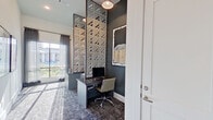 This single-floor business center is thoughtfully designed to blend functionality with contemporary aesthetics. The space features multiple dedicated workstations, each equipped with desktop computers and ergonomic chairs, set against deep charcoal accent walls that create a focused, professional atmosphere. A striking geometric lattice wall partition — clad in mirrored triangular panels — serves as a dramatic architectural centerpiece, adding depth and visual interest while subtly dividing the space.

Patterned carpet tiles in muted gray and navy tones run throughout, providing a sophisticated foundation that complements the overall design palette. Large windows dressed with sheer, dark-toned curtains allow abundant natural light to filter in, offering views of an outdoor pool area and surrounding landscaping. Framed abstract artwork adorns the walls throughout, reinforcing the space's polished, curated character.

The layout flows openly from a well-lit corridor — featuring soaring ceilings and additional framed art — into the individual work alcoves, creating a seamless transition between communal and focused areas. A printer station with additional counter space adds to the practical utility of the room. Clean white walls, crisp white trim, and recessed lighting complete the refined, modern look of this well-appointed workspace.