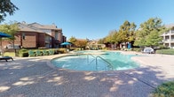 Located at 3620 Huffines Blvd in Carrollton, Texas, this well-appointed outdoor amenity space offers a range of recreational features set within a lushly landscaped environment. The centerpiece is a generously sized, freeform resort-style swimming pool with a connected wading or spa area, both surrounded by a textured concrete pool deck that provides ample space for lounging. Colorful Adirondack-style chairs and reclining sun loungers are positioned throughout the deck, inviting relaxation under the Texas sky. A striking white pergola serves as a shaded gathering area, furnished with round tables and vibrant blue and green seating, creating an ideal outdoor social hub. The space also features a fully equipped built-in outdoor kitchen, constructed with a stacked stone surround and topped with granite-style counters. The kitchen includes a large stainless steel gas grill and additional storage compartments, perfect for outdoor entertaining. Mature trees and manicured hedgerows frame the entire area, providing natural shade and privacy. The surrounding multi-story brick residential buildings are visible in the background, contributing a sense of community to the setting. The overall ambiance is bright, open, and inviting, with thoughtful landscaping and well-maintained amenities throughout.