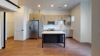 Located at 1050 Slater Rd in Durham, NC, this single-floor residence offers a thoughtfully designed layout with modern finishes and a bright, clean aesthetic throughout. The open-concept kitchen serves as a central highlight, equipped with stainless steel appliances including a range, microwave, dishwasher, and refrigerator with water dispenser. Light wood-toned cabinetry with matte black hardware, white quartz countertops, a subway tile backsplash, and a central island with a contrasting dark base provide both style and functionality. Warm hardwood-style flooring flows seamlessly through the kitchen and into the living area, which features crisp white walls, a ceiling fan with integrated lighting, and a window with blinds that invites natural light. Access to an outdoor balcony extends the living space further. The bathroom is finished with marble-look tile surrounds on the tub and shower combination, complemented by a curved glass shower door, a wood-toned vanity with white countertop, and coordinating hardwood-style flooring. Recessed lighting and consistent modern hardware details unify the overall design, creating a cohesive and contemporary living environment.
