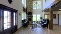 This single-floor home makes an immediate impression with its dramatic architectural design and thoughtfully curated interiors. The centerpiece of the living area is a commanding floor-to-ceiling stacked stone fireplace, which anchors the space and draws the eye upward toward the soaring double-height ceilings. A ceiling fan with a warm light fixture hangs above, adding both function and charm to the grand vertical space.

Large windows flank the fireplace and line the walls, flooding the interior with abundant natural light while offering sweeping views of the verdant landscape just beyond. The seamless connection between the indoors and the outdoors creates a bright, airy ambiance throughout.

The living room is furnished with elegant dark leather wingback chairs and a cowhide area rug atop rich wood-look tile flooring, blending rustic character with refined style. Exposed wooden beams accent the upper level, reinforcing the warm, ranch-inspired aesthetic that runs throughout the home.

Decorative wall sconces and tasteful art pieces add layered personality to the crisp white walls. A glimpse into adjacent spaces reveals an equally inviting interior, suggesting a well-connected, open floor plan designed for comfortable everyday living and effortless entertaining.