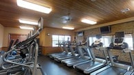This fitness center offers a comprehensive and inviting environment for residents or guests seeking a quality workout experience. The space is defined by its warm aesthetic, featuring a distinctive wood-plank ceiling that lends a lodge-like character, complemented by recessed flush-mount lighting that keeps the room bright and energetic throughout the day.

The cardio area is the centerpiece of the facility, housing a row of modern treadmills and additional cardio equipment such as ellipticals or stair climbers along the perimeter. Multiple wall-mounted flat-screen televisions are positioned throughout the room, ensuring entertainment is visible from virtually any machine.

Natural light filters in through large windows, creating an airy and open atmosphere that enhances the overall workout environment. A textured stone accent wall adds an additional layer of visual interest and architectural character to the space.

The flooring consists of dark rubber fitness flooring, which is both durable and practical for high-traffic exercise use. The warm earth-tone walls, combined with the natural wood ceiling and stone detailing, give the fitness center a distinctive, upscale feel that sets it apart from a standard gym environment. Overall, the space is thoughtfully designed to balance functionality with comfort.