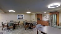 This single-floor communal space is designed to accommodate both casual gatherings and functional everyday use. The room is finished with neutral-toned walls and a dark ceiling that lends a contemporary contrast, complemented by a mix of recessed lighting and circular pendant fixtures that provide balanced, even illumination throughout.

The dining area serves as the focal point of the space, featuring multiple rectangular tables surrounded by upholstered chairs in a muted floral pattern. The seating arrangement is flexible, allowing the tables to host smaller groups independently or be used together for larger gatherings. Patterned low-pile carpeting in grey tones adds texture and visual interest underfoot.

Along one wall, a built-in kitchenette offers practical amenities, including warm medium-toned wood cabinetry, a stainless steel microwave, and a sink with counter space — well-suited for light food preparation or refreshment service. Decorative wall art, including a framed landscape photograph and an abstract metal wall accent, add personality and warmth to the room. The overall ambiance is clean, functional, and welcoming, reflecting a well-appointed shared common area.