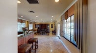 This single-floor space presents a thoughtfully designed interior that blends classic elegance with functional comfort. The entrance foyer immediately commands attention with its soaring vaulted ceiling, a decorative medallion inlaid into the polished travertine tile floor, and a striking arched window flanked by French doors that flood the space with natural light. A reception desk and informational displays suggest a welcoming, professional atmosphere.

The kitchenette area is well-appointed with rich cherry wood cabinetry featuring raised-panel detailing, granite countertops, a central island with bar-height seating for several guests, and essential appliances including a microwave, refrigerator, and small appliances. The space is brightened by large windows with horizontal blinds and decorative valances.

Adjacent to the kitchenette, the lounge and gathering area offers a warm, residential ambiance. It features a large, dark wood entertainment and display unit, plush upholstered sofas with botanical-patterned fabric, ornate carved wood dining chairs, and a vibrant traditional area rug anchoring the seating arrangement. Recessed lighting and crown molding throughout contribute to the refined aesthetic. The warm golden-toned walls and abundance of windows with outdoor views complete the inviting character of this well-appointed space.