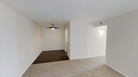This single-story space presents a straightforward, functional layout finished in a neutral color palette throughout. The interior is characterized by crisp white walls and white-painted ceilings that help reflect light and contribute to an open, airy feel. The living area is finished with light beige carpet, while an adjoining room features darker, brown-toned carpet, creating a subtle visual distinction between the two spaces. A partial wall divides the areas, adding a degree of architectural definition without fully closing off the floor plan. A ceiling fan with a brushed nickel finish is installed in the inner room, providing both air circulation and a modest lighting element. Recessed ventilation is visible in the ceiling, indicating a central HVAC system. White baseboards run along the perimeter of the rooms, offering a clean contrast against the flooring. The walls are smooth and unadorned, providing a blank canvas ready for personalization. The overall ambiance is calm and understated, with a move-in-ready condition that emphasizes simplicity and practicality throughout the space.