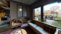 The Elysian's single-floor lobby and common area showcases a sophisticated industrial-chic aesthetic that blends raw architectural elements with refined furnishings. Polished concrete floors anchor the space throughout, while exposed overhead ductwork and piping lend an urban, loft-inspired character to the interior. Upon entry through the glass-paneled front doors — branded with The Elysian's distinctive logo — guests are welcomed into an open reception lounge featuring an ornate geometric chandelier, textured accent walls, and a curated arrangement of contemporary armchairs and settees that invite conversation and relaxation. The layout flows naturally into a comfortable seating nook lined with large windows overlooking an outdoor courtyard, where warm-toned leather sofas and richly patterned area rugs create an inviting retreat filled with natural light. Adjacent to this area, a well-appointed refreshment station offers a coffee bar with cabinetry, a sink, and beverage equipment, complemented by a wall-mounted television, pendant lighting, and a built-in dining banquette — ideal for casual gatherings. A darker, more intimate lounge zone features a sleek media wall with a television, additional seating, and subtle brick accents. Together, these areas create a cohesive, amenity-rich environment that balances industrial design with comfortable, welcoming warmth.