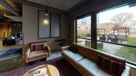 The Elysian's single-floor lobby and common area showcases a sophisticated industrial-chic aesthetic that blends raw architectural elements with refined furnishings. Polished concrete floors anchor the space throughout, while exposed overhead ductwork and piping lend an urban, loft-inspired character to the interior. Upon entry through the glass-paneled front doors — branded with The Elysian's distinctive logo — guests are welcomed into an open reception lounge featuring an ornate geometric chandelier, textured accent walls, and a curated arrangement of contemporary armchairs and settees that invite conversation and relaxation. The layout flows naturally into a comfortable seating nook lined with large windows overlooking an outdoor courtyard, where warm-toned leather sofas and richly patterned area rugs create an inviting retreat filled with natural light. Adjacent to this area, a well-appointed refreshment station offers a coffee bar with cabinetry, a sink, and beverage equipment, complemented by a wall-mounted television, pendant lighting, and a built-in dining banquette — ideal for casual gatherings. A darker, more intimate lounge zone features a sleek media wall with a television, additional seating, and subtle brick accents. Together, these areas create a cohesive, amenity-rich environment that balances industrial design with comfortable, welcoming warmth.