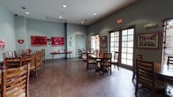 This single-floor common room offers a versatile and welcoming gathering space, thoughtfully designed to accommodate a variety of events and activities. The room features rich, dark hardwood flooring that anchors the space and complements the cool gray-toned walls, creating a warm yet contemporary ambiance throughout.

Recessed lighting is evenly distributed across the ceiling, providing consistent illumination that is supplemented by generous natural light streaming through large French doors along one wall. These glass-paned doors offer views of a lush outdoor area and provide easy access to the exterior, enhancing the room's open and airy feel.

The space is furnished with a mix of round dining tables and an assortment of wooden chairs, allowing for flexible seating configurations suited to both intimate gatherings and larger group events. A folding table along one wall provides additional surface space for serving or displays.

Seasonal decorations, currently themed in red and pink, adorn the walls alongside framed artwork and photographs, adding a festive, personalized character to the room. A standard interior door provides access to adjacent areas, and a utility panel is discretely mounted on the wall. Overall, the room is well-suited for communal dining, celebrations, and organized activities.