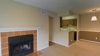 This single-floor space combines a comfortable living area with a functional kitchen in an open layout. The living room is anchored by a wood-burning fireplace framed in warm terracotta-toned tile with a clean white mantel, adding a focal point to the room. Neutral beige carpeting covers the living area floor, contributing to the warm, understated ambiance throughout. Smooth, light-colored walls are complemented by vaulted ceiling lines, lending a sense of openness to the interior.

The kitchen is positioned adjacent to the living area and is defined by a peninsula counter that serves as a natural divider between the two spaces. Medium-toned wood cabinetry lines the walls above, providing ample storage, while a sink is visible along the counter. A black appliance is situated beneath the counter, likely a dishwasher. Tile flooring in the kitchen area contrasts with the carpeting of the living space, clearly delineating the two zones. A ceiling-mounted light fixture brightens the kitchen, and a ventilation grille is visible in the wall, indicating a central HVAC system throughout the unit.