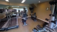 This fitness center offers a practical and functional workout environment designed to support a range of exercise routines. The space is furnished with multiple cardio machines, including treadmills, upright and recumbent stationary bikes, and an elliptical trainer, providing users with ample options for cardiovascular exercise. A multi-station strength training machine is also available for resistance-based workouts. A wall of mirrors spans one side of the room, enhancing the sense of space and allowing users to monitor their form during exercise. A flat-screen television is mounted near the ceiling, offering entertainment to keep workouts engaging. The room features warm, neutral-toned walls and patterned carpeted flooring, contributing to a comfortable and inviting atmosphere. Recessed and panel lighting ensures the space is well-lit throughout. A window along the mirrored wall allows for natural light to filter in, adding brightness to the room. The overall layout is thoughtfully organized to allow for ease of movement between equipment, making this fitness center a convenient and comprehensive amenity for health-conscious users.
