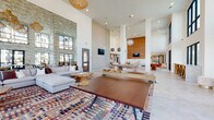 Located in Sarasota, FL, this expansive single-floor clubhouse showcases a bold, contemporary design defined by soaring double-height ceilings and an abundance of natural light streaming through large, black-framed floor-to-ceiling windows. The open-concept layout flows seamlessly across distinct functional zones, all unified by light-toned herringbone and hexagonal tile flooring.

The main lounge area is anchored by an oversized sectional sofa, colorful woven area rugs, and a sculptural wood coffee table, complemented by woven rattan pendant chandeliers and a dramatic textured accent wall. A game area features a ping-pong table and unique wooden seating, opening directly to outdoor views of palm trees and a serene waterfront.

A dedicated dining and bar area includes counter-height seating along black-framed glass walls, offering an airy, café-like atmosphere with generous views of the surrounding landscape. The full kitchen is outfitted with stainless steel appliances, terrazzo-style stone countertops, herringbone tile backsplash, and a mix of open shelving and cabinetry in warm wood and muted gray tones.

Throughout the space, warm globe wall sconces and sculptural pendant lighting add an inviting ambiance, while white structural columns and expansive glazing reinforce the sense of openness. Outdoor views of the waterfront and palm-lined grounds are visible from nearly every corner of this thoughtfully designed communal space.