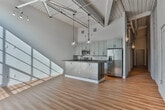 Located at 710 Greenville St in LaGrange, Georgia, this single-floor loft-style unit showcases a thoughtful blend of industrial character and contemporary design. The space retains its original architectural identity through white-painted exposed brick walls, visible structural beams, and open ceiling details including exposed copper piping and ductwork — all hallmarks of a carefully renovated historic building.

The open main living area features warm hardwood flooring, soaring ceilings, and a large industrial-style multi-pane window that floods the space with natural light. A ceiling fan and track lighting provide both comfort and ambiance. The kitchen is centrally positioned and defined by a peninsula with dark countertops offering bar seating, gray shaker cabinetry, a tile backsplash, stainless steel appliances, and pendant globe lighting that adds a modern touch overhead.

A long corridor lined with exposed brick leads to additional rooms, including a well-proportioned private room with tall ceilings and white panel doors. The bathroom features warm hardwood flooring, a dark-countered vanity with cabinetry, a round mirror, and clean, neutral finishes. A sliding barn-style door adds a functional, stylish detail. Throughout, the space balances raw industrial elements with polished modern upgrades.