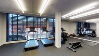 This single-floor fitness center is designed to provide a comprehensive workout environment with a clean, modern aesthetic. The space is outfitted with multiple treadmills and stationary cycling equipment positioned along a wall of large, horizontal-slatted windows that allow generous natural light to filter in while offering views of the surrounding exterior — creating an energizing atmosphere for exercise.

Structural columns punctuate the layout, subtly defining zones within the open floor plan. The flooring consists of durable, low-pile carpet well-suited for gym use, while the drop ceiling features recessed linear lighting that ensures bright, even illumination throughout. A mirrored wall on one side visually expands the space and supports stretching and form-checking during workouts.

The fitness area also includes a weight bench and a collection of medicine balls stored neatly in a rack, catering to both cardio and strength training needs. The overall ambiance is functional and well-maintained, with a layout that encourages efficient movement between equipment. The combination of natural light, mirrors, and modern equipment makes this fitness center a practical and inviting space for a wide range of exercise routines.