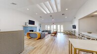 This thoughtfully designed single-floor communal space offers a generous, open layout with a bright, contemporary aesthetic throughout. The expansive living room serves as the centerpiece, featuring striking skylights and recessed lighting that flood the interior with natural light. Light wood-look flooring flows seamlessly across the entire space, enhancing its airy, cohesive feel. A standout blue mosaic tile fireplace anchors the main seating area, which is complemented by a wall-mounted TV, large-scale abstract artwork, and a curated arrangement of mid-century modern and contemporary furnishings. Multiple seating zones provide flexible gathering areas, while a bar-height counter with gold-accented stools offers additional space for socializing. The kitchen area is visually distinctive, featuring white shaker cabinetry with brushed gold hardware, a sleek quartz countertop, a built-in wine refrigerator, and floor-to-ceiling patterned blue-and-white encaustic-style tile that creates a bold, decorative backdrop. Rustic wood floating shelves add warmth and display space. The entrance corridor transitions naturally into the living area and features full-length glass French doors that open to a private outdoor terrace, offering views of a pool and the surrounding Mediterranean-style architecture, seamlessly connecting the interior to the outdoors.