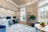 Located at 680 SC-160 in Fort Mill, South Carolina, this thoughtfully designed office space offers a refined and functional environment ideal for productivity and collaboration. The single-room layout is elevated by a striking tray ceiling with recessed lighting and a decorative sputnik chandelier that serves as a glamorous focal point overhead.

The walls feature a sophisticated combination of textured wallcovering above and classic wainscoting panels below in a soft blue-gray tone, creating a polished, layered aesthetic throughout. Plush, patterned carpet in muted blue and gray tones adds warmth and comfort underfoot.

A built-in workstation with dual monitors and a printer is positioned along one wall, supported by ample cabinetry beneath, providing a well-equipped workspace for multiple users. Adjacent to the workstation, a tall brass-framed open shelving unit adds both storage and decorative appeal.

Large windows along two walls flood the space with abundant natural light while offering views of the surrounding landscape. A cozy seating nook near the windows features a round pedestal table with bistro-style chairs — perfect for casual meetings or consultations. On the opposite side, a pair of plush blue velvet armchairs flank a small side table beneath a wall-mounted flat-screen television, creating an inviting lounge area. The overall ambiance is professional yet welcoming, balancing function with refined interior design.