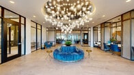 Located at 5905 Caravan Ct in Orlando, Florida, this sophisticated single-floor lobby space offers a striking first impression through its contemporary architectural design and carefully curated interior details. The centerpiece of the space is a spectacular large-scale circular chandelier composed of dozens of globe pendant lights suspended at varying heights, creating a dramatic focal point that anchors the open, airy main area below. Large-format stone tile flooring in soft neutral tones extends throughout, lending a polished and cohesive aesthetic. A circular, tufted velvet seating piece in bold blue sits beneath the chandelier, complemented by sleek brass accent tables that add warmth and sophistication. Floor-to-ceiling glass walls and expansive windows flood the interior with natural light while offering views of the surrounding outdoor landscape, including palm trees that hint at the Florida setting. Black steel-framed glass partitions divide the space into distinct functional zones, including private workstation alcoves adorned with an eye-catching marbled wall mural in gold and neutral tones. A dedicated reception area features a built-in wood and white countertop desk beneath geometric black metal pendant fixtures. The overall ambiance is refined, modern, and welcoming, balancing open communal areas with more intimate, purpose-driven spaces.