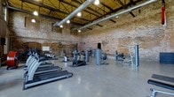 This fitness facility showcases a striking industrial aesthetic defined by soaring ceilings with exposed steel trusses and ductwork, worn brick walls, and polished concrete flooring. The open, warehouse-style layout provides an expansive workout environment with ample space between equipment stations. Overhead pendant lighting illuminates the floor, creating a functional and energetic atmosphere.

The cardio area features a row of True treadmills along one side, offering dedicated space for aerobic training. The strength and functional training zones include a variety of resistance machines, a cable system, and a weight rack, giving users access to a comprehensive selection of equipment. The open floor plan allows for easy circulation and flexible use of the space.

The raw, industrial character of the brick walls and structural ceiling elements contrasts with the modern fitness equipment, lending the facility a distinctive, urban character. The high ceilings contribute to an open, airy ambiance, making the space feel both functional and visually compelling for an engaging workout experience.