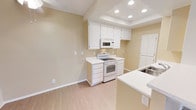 This single-story space presents a clean, functional design centered around an open kitchen and adjoining living area. The kitchen is equipped with white cabinetry that extends to the ceiling, offering generous storage capacity. White appliances, including an electric range and an over-the-range microwave, complement the light-toned countertops, which also form a peninsula that serves as a natural divider between the kitchen and the adjacent living space. A stainless steel sink is integrated into the peninsula, adding a practical workspace. A distinctive tray ceiling with recessed lighting sits above the kitchen, providing focused illumination while adding architectural interest. The adjacent living area features warm beige walls and light wood-tone flooring that extends throughout, contributing to a cohesive and airy atmosphere. A ceiling fan with an integrated light fixture serves the living area, enhancing comfort and air circulation. Clean white baseboards frame the space consistently, and multiple electrical outlets are positioned conveniently throughout. The overall palette of soft neutrals and white accents creates a bright, welcoming environment that feels open and well-maintained.
