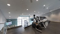 The Haven's fitness center is a well-appointed, single-floor facility designed to support an active lifestyle. The space is outfitted with a comprehensive selection of modern cardio equipment, including stair climbers, ellipticals, and treadmills, all arranged along one side of the room to maximize open floor space. The ceiling features an array of recessed lighting that ensures bright, even illumination throughout the workout area, complementing the natural light that filters in through large windows along the far wall. These windows not only brighten the space but also provide views to the exterior, creating an open and airy atmosphere during workouts. The interior design incorporates a neutral color palette accented by teal-toned wall sections, lending the space a clean and contemporary feel. Glass railings line the upper-level overlook, reinforcing the modern aesthetic while maintaining sightlines across the facility. A wall-mounted television is positioned for visibility from the cardio equipment, adding to the amenity's functional appeal. The flooring is a durable, dark-toned material well-suited for high-traffic fitness use. Overall, the fitness center at The Haven combines practical equipment, thoughtful design, and an inviting environment.