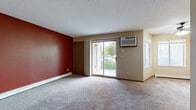 This single-floor unit offers a practical and functional layout spread across several distinct areas. The spacious living room serves as the heart of the space, featuring plush wall-to-wall carpeting, a bold deep-red accent wall, and a textured ceiling. Natural light fills the room through multiple windows and a sliding glass door that opens directly to an outdoor green area, providing pleasant views of the surrounding landscape. A wall-mounted air conditioning unit and baseboard heating ensure year-round comfort. A ceiling fan adds additional airflow to the bright, windowed corner of the living area.

The kitchen is efficiently designed with warm oak cabinetry, laminate countertops, a double sink, and tile flooring in a neutral stone pattern. White appliances, including a refrigerator, electric range, and dishwasher, are neatly integrated into the layout.

The bedrooms are carpeted and finished in neutral tones, each offering a calm, understated atmosphere. One bedroom features a window with a baseboard heater beneath it, allowing ample natural light. The bathroom is accessed via a tiled hallway and includes a vanity with wood cabinetry, a toilet, and a tub-shower combination. Wood-toned doors and trim are consistent throughout, tying the overall aesthetic of the unit together cohesively.